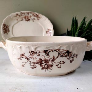 May include: Cream-coloured antique serving bowl with brown floral and scrollwork design. The bowl has two handles and a matching plate in the background. The set is placed on a white surface with a green plant behind.