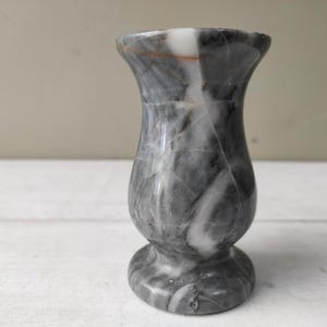 Op de afbeelding: Een kleine, decoratieve vaas van grijs en wit marmer. De vaas heeft een uitlopende bovenkant, een afgeronde body en een cirkelvormige basis. Het marmer heeft een natuurlijk, wervelend patroon.