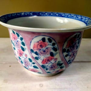 May include: A ceramic bowl with a white interior and a pink exterior decorated with floral patterns in pink, teal, and white. The rim is white with a blue geometric design. The bowl is likely for decorative purposes.