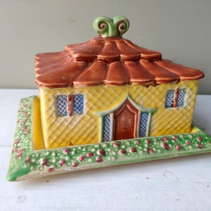 May include: A ceramic house-shaped butter dish. The roof is brown, the walls are yellow, and the base is green with red accents. The house has blue-framed windows and a red door. The top has a green swirl design.