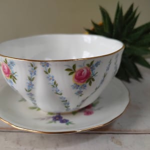 Könnte beinhalten: Eine weiße Porzellan-Teetasse und -Untertasse mit einem Blumenmuster. Tasse und Untertasse haben einen goldenen Rand und zeigen blaue und rosa Blumen.