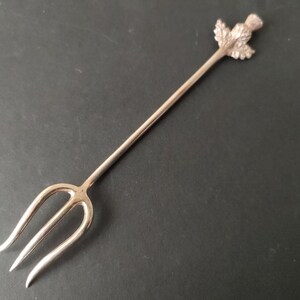 May include: Silver-coloured serving fork with a three-pronged end and a thistle design at the top. The fork is made of metal and is set against a dark background. The thistle design is detailed and adds a decorative element.