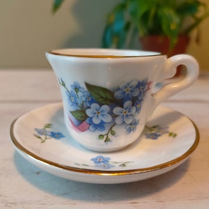 May include: A white teacup and saucer set with a gold trim and a floral design featuring blue forget-me-not flowers.
