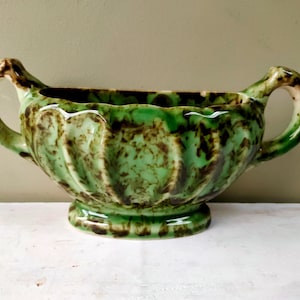 Op de afbeelding: Een vintage keramische plantenbak met een groen en bruin gevlekt glazuur. De plantenbak heeft een geschulpte rand, twee gebogen handvatten en een voetstuk. Dit decoratieve stuk is geschikt voor binnen of buiten.