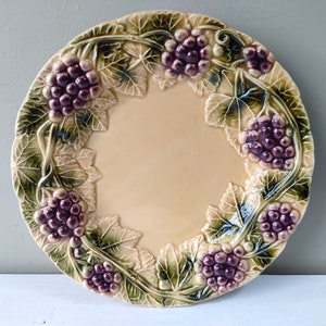 Puede incluir: Un plato redondo de cerámica color crema con un diseño en relieve de uvas y hojas alrededor del borde. Las uvas son moradas y las hojas verdes. El plato tiene un centro liso y sin adornos.