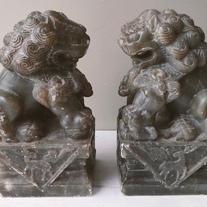 May include: A pair of gray stone foo dog statues with intricate details. The statues are sitting on square bases with carvings.