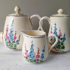 Pode incluir: Um conjunto de chá creme com bule, cremeira e cafeteira. Cada peça é adornada com um padrão floral em tons de rosa e azul, com detalhes dourados. Ideal para servir chá ou café.
