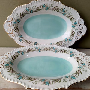 Op de afbeelding: Twee ovale serveerschalen met blauwe en witte bloemenmotieven en gouden randjes. De schalen hebben een lichtblauwe glazuur en zijn perfect om hapjes of voorgerechten op te serveren.