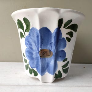 Op de afbeelding: Witte keramische bloempot met een blauw bloemmotief en groene bladeren. De bloem is in het midden van de pot geschilderd. De pot heeft een geschulpte rand en een licht uitlopende vorm, geschikt voor binnen of buiten.