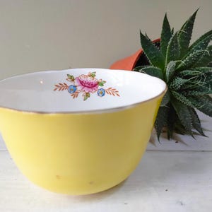 May include: A yellow teacup with a white rim and a floral design featuring a pink rose and blue flowers.