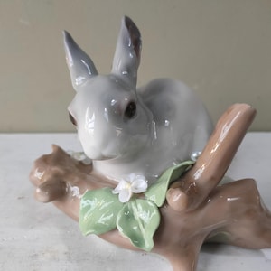 Może przedstawiać: Porcelanowa figurka szarego królika siedzącego na gałęzi z białymi kwiatami. Królik siedzi na gałęzi z zielonymi liśćmi i białymi kwiatami.
