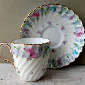 May include: A white teacup and saucer with a floral pattern in shades of pink, blue, and green. The cup has a fluted rim and a swirling design. The saucer has a scalloped edge.