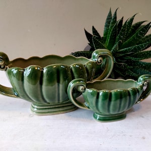 May include: Two green ceramic planters with scalloped edges and curved handles. The larger planter is approximately 12 inches long, and the smaller one is about 6 inches long. Both have a glossy finish and are placed on a white surface.