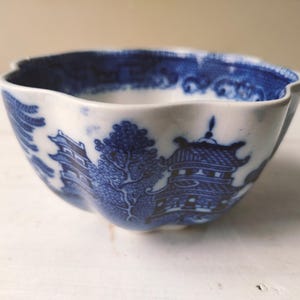 May include: A white and blue ceramic bowl with a scalloped rim. The bowl features a blue Willow pattern design with pagodas, trees, and bridges. The bowl is likely vintage or antique.