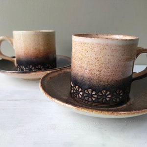 May include: Two brown and beige ceramic coffee cups with matching saucers. The cups have a dark brown band with a decorative floral pattern around the top.