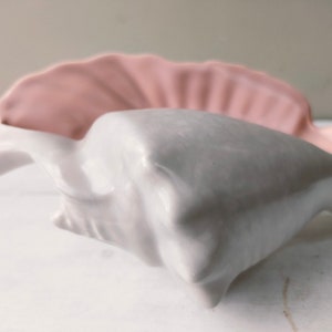 Op de afbeelding: Een set van twee keramische kommen in de vorm van een schelp. De ene kom is wit en de andere is roze.