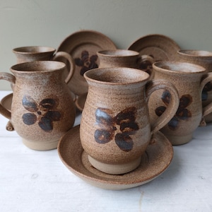 Puede incluir: Juego de seis tazas de gres marrón con un diseño floral. Cada taza tiene un platillo a juego. Las tazas y los platillos tienen un aspecto rústico y hecho a mano.