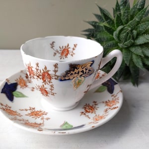 May include: White teacup and saucer set with orange floral and blue leaf designs. The cup and saucer have a scalloped edge. The teacup has a gold and brown design on the side. A decorative item for the kitchen.