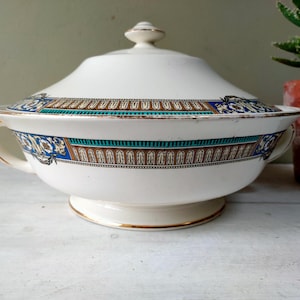 Puede incluir: Fuente de cerámica blanca con tapa y una banda decorativa con diseños azules, turquesas y marrones. El plato tiene dos asas y un borde dorado. Un pequeño pomo redondo se encuentra en la parte superior de la tapa.