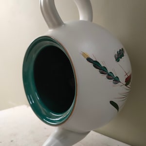 May include: White ceramic teapot with a green rim and a floral design featuring wheat stalks. The teapot has a handle and a spout.