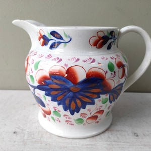 Peut inclure: Un pichet en céramique blanche avec un motif floral dans les tons de bleu, rouge, vert et or. Le pichet a un corps arrondi et une anse.