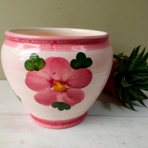 Peut inclure: Pot de fleur en céramique avec un motif rose et blanc. Le pot présente une grande fleur rose avec un centre jaune et des feuilles vertes. Le bord et la base sont également peints en rose. Une plante verte est visible en arrière-plan.
