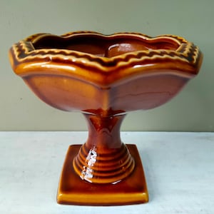 Peut inclure: Un bol à piédestal en céramique vintage dans les tons de marron et d'orange. Le bol a un bord festonné et repose sur une base carrée. Le piédestal a un design nervuré. Cette pièce décorative convient pour exposer des fruits ou autres.