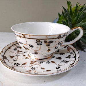 May include: A white teacup and saucer with a black and gold floral design. The cup has a delicate handle and a wide, shallow bowl. The saucer has a matching design and a scalloped edge.