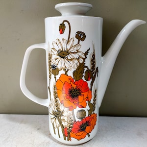 Może przedstawiać: Biały ceramiczny dzbanek do kawy z kwiatowym wzorem przedstawiającym pomarańczowe maki, białe stokrotki i kłosy pszenicy. Dzbanek ma pokrywkę i dzióbek.