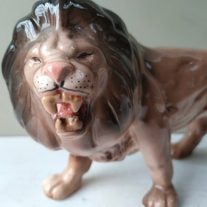 Puede incluir: Una figurita de cerámica de un león rugiente con una melena marrón y negra. El león está de pie sobre sus cuatro patas con la boca abierta y los dientes visibles.