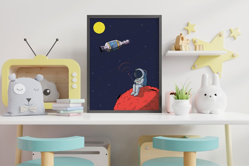 Puede incluir: Ilustraci&oacute;n enmarcada de un astronauta sentado en un planeta rojo, sosteniendo un dispositivo con l&iacute;neas de se&ntilde;al. Un sat&eacute;lite y una luna amarilla est&aacute;n en el cielo azul oscuro estrellado. La obra de arte se exhibe sobre una mesa blanca.