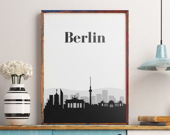 May include: A framed print featuring the word "Berlin" above a black and gray silhouette of the Berlin skyline. The print is in a distressed wooden frame and is displayed on a blue shelf with a vase of white flowers.