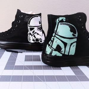 May include: A pair of black high-top sneakers with custom painted Star Wars characters. One shoe features a white and black Stormtrooper, while the other shoe features a green and black Boba Fett.