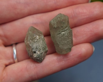 May include: Two rough-cut, light brown quartz crystals are held in a hand. The crystals have a natural, unpolished appearance, with visible textures and variations in color. The background is a blurred mix of green and blue.