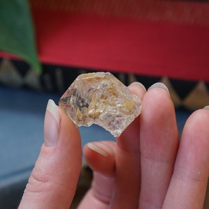 Puede incluir: Un cristal de cuarzo transparente con inclusiones doradas. El cristal está facetado y tiene una textura rugosa y natural.