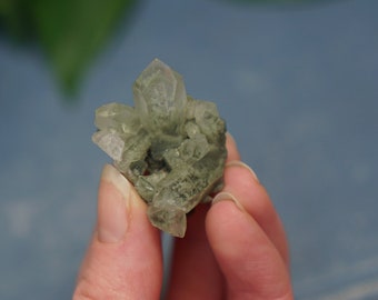 May include: A small, clear quartz crystal cluster with green inclusions. The crystal is being held in a hand.