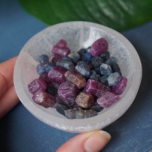 Peut inclure: Un petit bol rond blanc rempli d'un mélange de pierres précieuses brutes, non taillées, dans des tons de violet profond, de bleu et de gris. Le bol est tenu au-dessus d'un livre bleu, avec une feuille verte en arrière-plan.