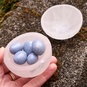 May include: A selenite crystal bowl with a smooth, white surface holds a collection of light blue, smooth, polished stones. The bowl is in the palm of a hand.