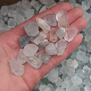 May include: A hand holding a pile of small, light blue aquamarine crystals. The crystals are translucent and have a variety of shapes and sizes.
