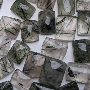 May include: A collection of green and clear quartz cabochons with black inclusions. The cabochons are all rectangular and have a smooth, polished finish.