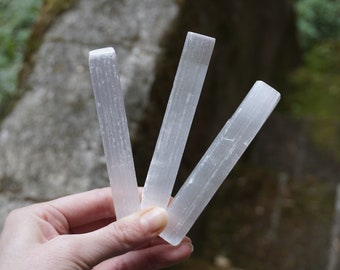 May include: Three white selenite wands, also known as selenite sticks, are held in a hand. The wands are smooth and have a natural, striated texture.