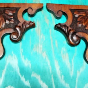 May include: A pair of L-shaped wooden corbels featuring intricate carved leaf patterns and scrollwork details. The wood has a medium brown finish with darker stained recessed areas, showcasing a symmetrical design with curved edges and sharp corners.