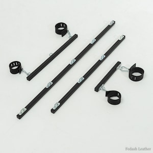 May include: An overhead shot shows a set of black restraints with metal hardware. The set includes four black bars with metal attachments and four black cuffs. The cuffs are connected to the bars with metal chains.