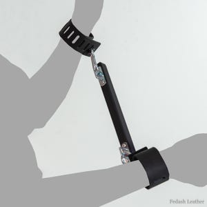 May include: Black leather wrist restraints connected by a black bar with metal hardware. The restraints have multiple slots for adjustment. The image is set against a plain white background.