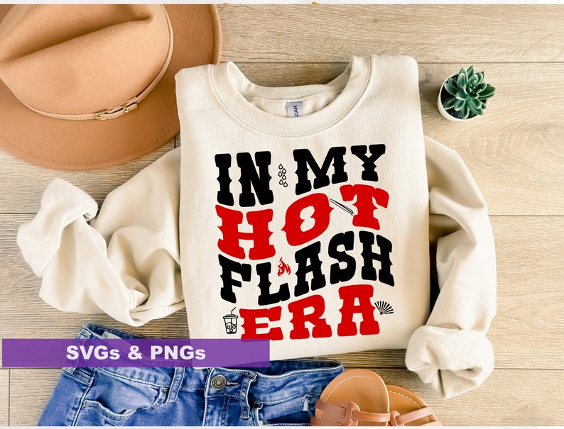 May include: A white sweatshirt with the text "IN MY HOT FLASH ERA" in black and red letters. The text is in a bold, blocky font. The sweatshirt is laid out on a wooden surface with a brown hat, a pair of sandals, and a small potted plant.