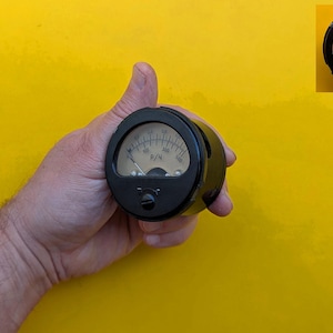 Puede incluir: Un medidor analógico redondo de color negro con una esfera beige y una aguja que apunta hacia la izquierda. El dispositivo presenta marcas negras y el texto cirílico P/Ч impreso en el centro. Tiene una carcasa circular negra y un pequeño botón.