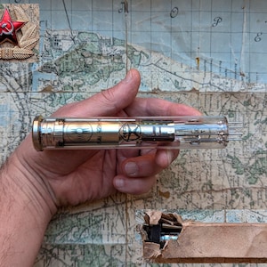Puede incluir: Un tubo de vacío de vidrio vintage con una base de metal plateado y un cuerpo de vidrio transparente. El tubo tiene una etiqueta roja y blanca con el texto "6Ж1П 142MH" y un símbolo en blanco y negro de una onda. El tubo está en una caja de cartón.