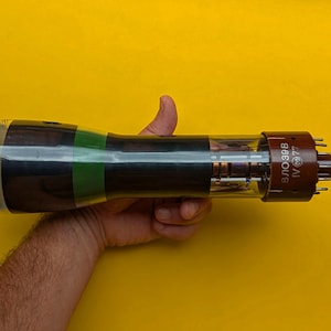 Può includere: Un tubo a vuoto cilindrico in vetro con una base marrone scuro che riporta il testo 8LO39B IV 77 e un logo circolare. Il tubo ha un'estremità anteriore ampia e arrotondata con una fascia verde e un corpo in vetro trasparente.