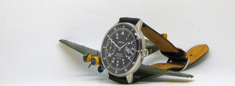 May include: A silver-cased wristwatch with a black face and white numerals, featuring a leather band. The watch is displayed on a small, olive-green model airplane with yellow accents. The watch face includes the text "SPECIAL FOR NAVIGATOR".