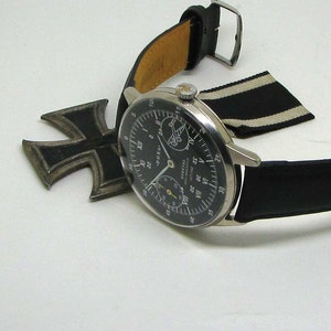 May include: A vintage wristwatch with a black leather band and a silver-colored case. The watch face is black with white and yellow markings. An Iron Cross medal and a black and white striped ribbon are also present.